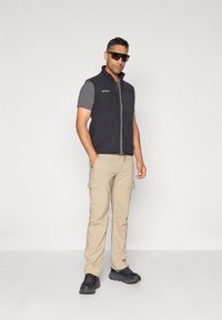 Crni fleece prsluk preko sive majice kratkih rukava, bež cargo hlače i crne sportske cipele. Model nosi tamne sunčane naočale.
