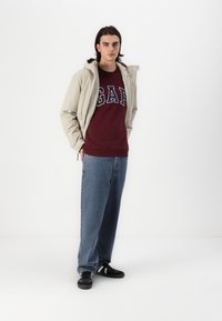 Beige hupparitakki maroon-colan sweatshirtin, jossa on "GAP"-logo. Yhdistetty sinisiin löysiin farkkuihin ja mustiin lenkkareihin. Yksinkertainen muotoilu, ei kuvioita.