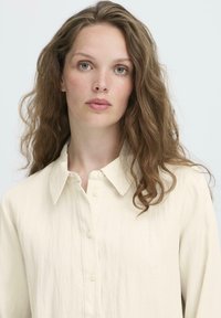 Woman with wavy light brown hair wears a cream-colored button-up shirt, looking directly ahead against a plain light background.