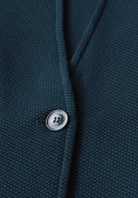 Texturerad mörk teal kofta med en notchad krage och en silverknapp med varumärket "StreetOne" synlig.