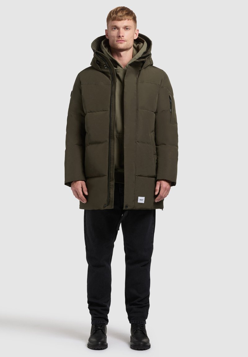 Maslinzasta pufasta jakna s kapuljačom, zatvaranjem na zip i bočnim džepovima, složena preko tamnog dukserice i crnih traperica, nosi se s crnim čizmama.