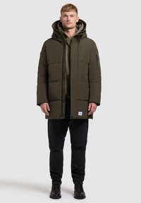 Gewatteerde groene parka-jas met ritssluiting, grote capuchon, zijzakken en een gestructureerde stof; gedragen over een donker hoodie en broek.