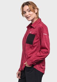 Rot kariertes Hemd mit langen Ärmeln, schwarzer Brusttasche, durchgehender Knopfleiste und Logo-Akzent am Arm. Leichte Textil mit glatter Oberfläche.