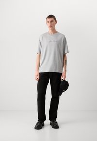 Gray cotton t-shirt with embroidered "Calvin Klein" logo, paired with black pants and a black cap. Model stands against a plain background.
