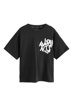 Niño de pie con las piernas separadas, vistiendo una camiseta negra extragrande con texto blanco, vaqueros gris oscuro y zapatillas blancas sobre fondo liso.