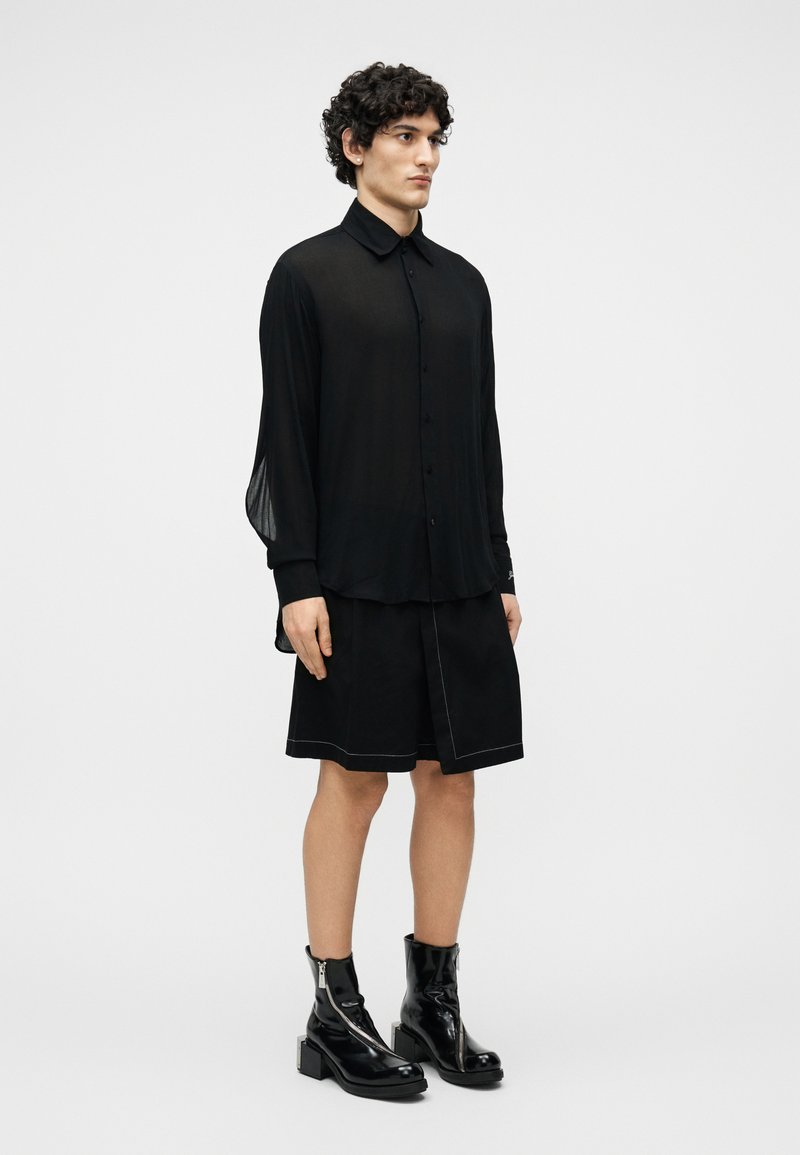 Young person with curly hair wearing a sheer black button-up shirt, black knee-length shorts, and shiny black ankle boots.