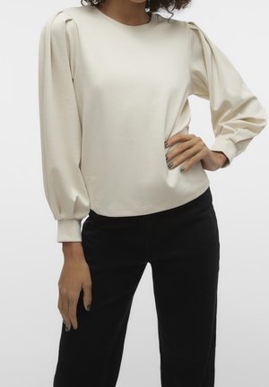 Kvinde iført en cremefarvet bluse med lange ærmer og puffede skuldre samt sorte bukser, stående med den ene hånd på hoften.