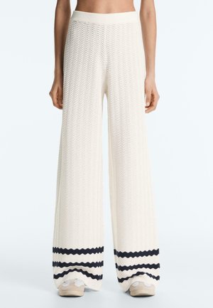 Nő fehér, bőszárú, texturált nadrágot visel fekete vízszintes csíkokkal a szegély közelében és bézs sportcipőt, egy egyszínű háttér előtt állva.