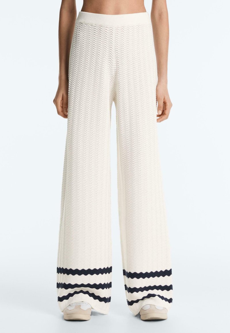 Femme portant un pantalon large blanc texturé avec des rayures horizontales noires près de l'ourlet et des baskets beiges, debout devant un fond uni.