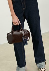 Femme portant un jean roulé bleu foncé et des chaussures à imprimé léopard, tenant un petit sac à main rectangulaire marron foncé avec un porte-clés en denim.