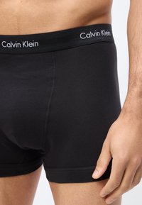 Mand iført sorte Calvin Klein boxershorts med synligt logo på linningen og afslappet hånd ved siden mod en enkel baggrund.