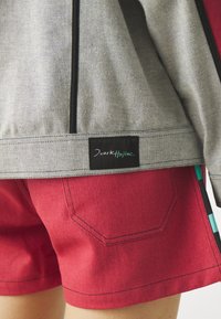 Gray denim jacket with black zipper, paired with red shorts featuring black stitching and teal accent detailing. Brand label displayed.