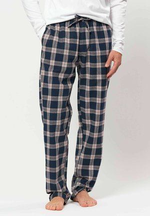 Homme portant un pantalon de pyjama à carreaux bleu marine et blanc avec une chemise blanche à manches longues, debout pieds nus sur un fond blanc.