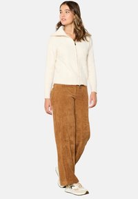 Kreminė kabelinė megzta zip-up kardiganas, derinamas su plačiomis rudomis aksominėmis kelnėmis ir šviesiai rudais sportiniais batais. Modelis stovi atsipalaidavęs, šiek tiek pasisukęs.