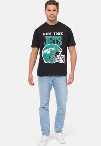 Černé bavlněné tričko s nápisem "NEW YORK JETS" v bílé a tyrkysové barvě, s grafikou fotbalové helmy. V kombinaci se světle modrými džíny.