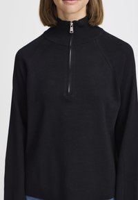 Pullover nero con colletto alto e chiusura a zip. Presenta maniche raglan e una texture a maglia liscia.