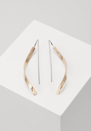 Par af guldfarvede snoede metaltrådøreringe med lange sølvkroge vist på en hvid geometrisk overflade.