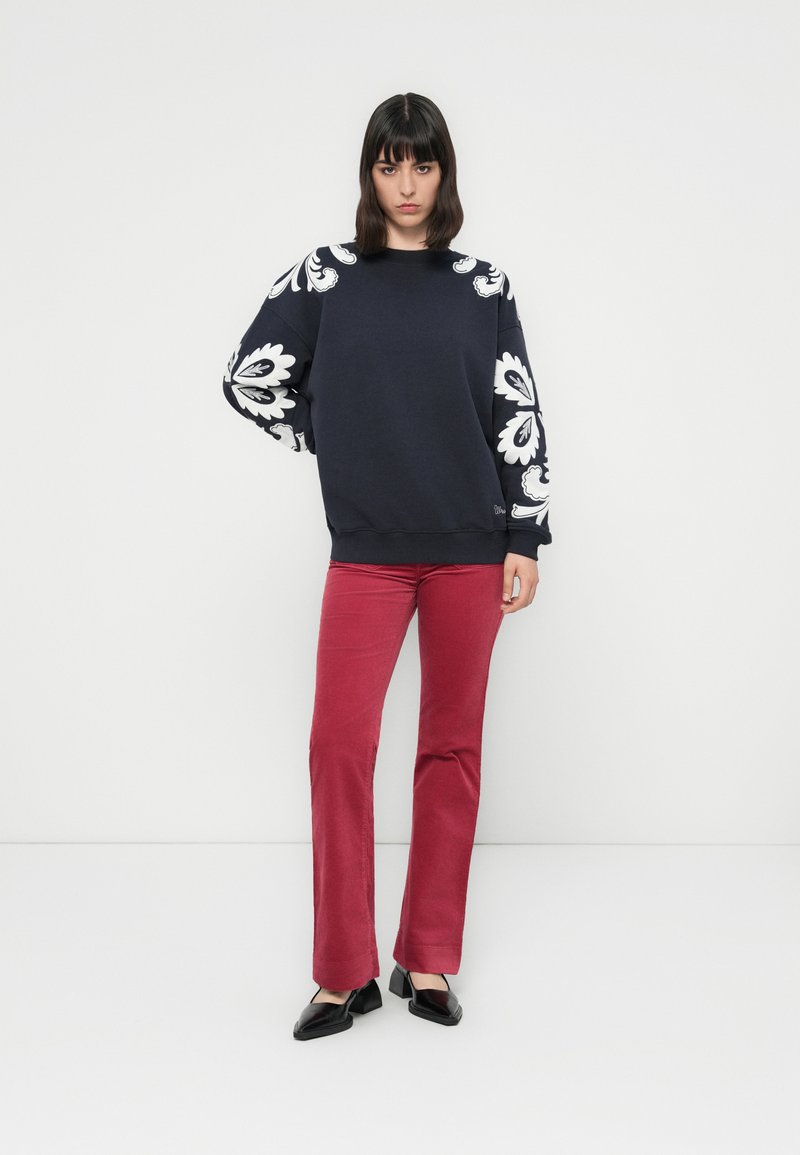 Marineblauwe sweatshirt met witte bloemenpatronen op de mouwen, gecombineerd met rode, strakke broek en zwarte platte schoenen. Eenvoudige halslijn en omgeslagen zomen.