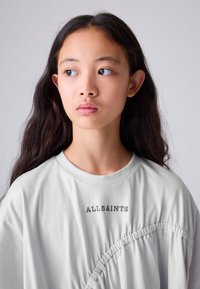Svijetlosiva oversize majica s naboranom detaljom i vezenim natpisom "ALLSAINTS" u tamnosivoj boji. Glatka tkanina i zaobljeni izrez oko vrata.