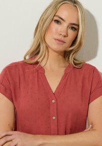 Blusa de manga corta en color coral con escote en V, que presenta pequeños puntos texturizados y cierre de botón, fabricada en una tela ligera.