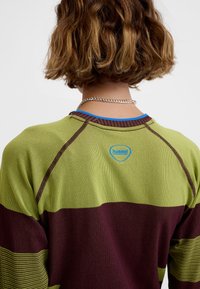 Kortærmet skjorte i grøn og burgunder, med ribstrikket tekstur, kontrasterende farveblokke og en blå logo ved nakken på bagsiden.