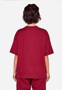 Burgundi színű, bő fazonú póló puha pamutból, kerek nyakkivágással és rövid ujjakkal, laza szabással és varrott szegély részletekkel.