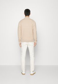 Maglione beige con texture e collo rotondo, visto da dietro. Abbinato a pantaloni chiari e sneakers bianche, su uno sfondo semplice.
