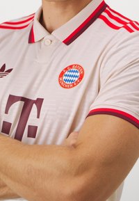 Lähedalt vaade heledale beežile FC Bayern Munichi jalgpallijerseysile, millel on punased ja tumepunased kraestriibid ning rinna peal logo, mida kannab käed ristis inimene.