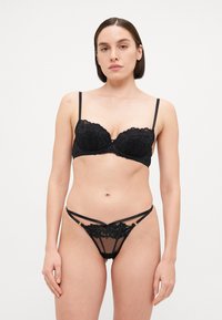 Ensemble de lingerie noir comprenant un soutien-gorge balconnet en dentelle avec des bords festonnés et un string avec une dentelle superposée et des accents de bretelles. Texture transparente présente.