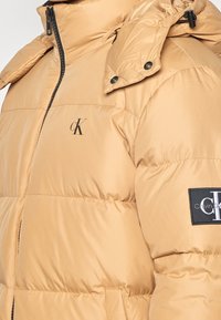 Veste matelassée beige avec fermeture éclair noire, capuche à boutons-pression, petit logo "cK" sur la poitrine et écusson Calvin Klein sur la manche gauche.