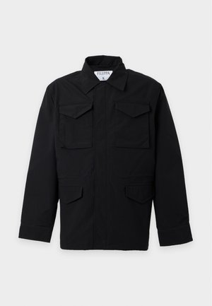 Homme debout portant une veste noire avec quatre poches avant, un pantalon large noir, des chaussures noires et une chemise blanche sous la veste.