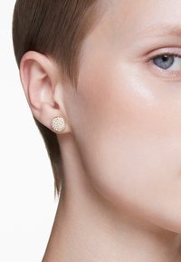 Brinco de ouro em forma redonda, apresentando um cluster de gemas transparentes que brilham contra um tom de pele suave e cabelo curto.