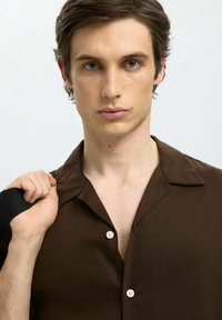 Brun kortärmad skjorta med krage, prydd med två vita knappar och en texturerad tyg. Modellen håller i en svart rem med ena handen.