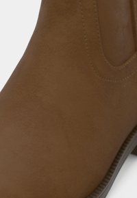 Καφέ ψεύτικο δερμάτινο μποτάκι αστραγάλου με λεία υφή και ελαστικές πλαϊνές πάνελ, διαθέτοντας χαμηλή ελαστική σόλα και διακριτική λεπτομέρεια ραφής.