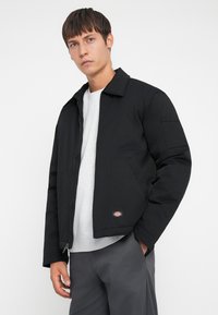 Black zip-up jacket with a collar, featuring a chest pocket and logo patch. Worn over a grey sweater and paired with grey pants.