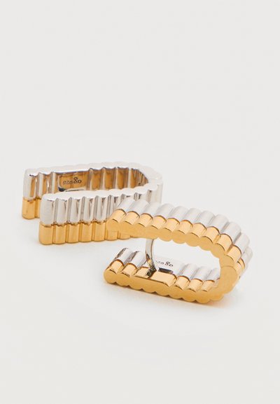 Δύο ανοιχτοί ορθογώνιοι δακτύλιοι με ραβδωτό σχεδιασμό, με εναλλασσόμενους ασημένιους και χρυσούς τόνους σε απλό φόντο.