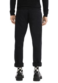 Man staat met ontspannen handen, gekleed in zwarte opgerolde broek, zwarte schoenen en zwart-witte geruit sokken tegen een witte achtergrond.
