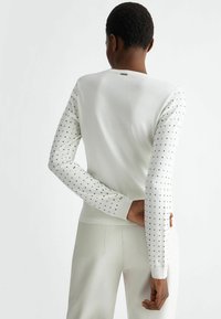 White sweater with long sleeves featuring metal studded accents along the sleeves. Soft knit material and a round neckline.