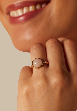 Main avec bague en or ornée d'une pierre rose ronde, touchant le menton près des lèvres souriantes et des dents sur fond neutre.