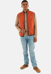 Gilet orange rembourré avec un col haut et une fermeture éclair, porté sur un pull crème. Un jean bleu clair et des chaussures brunes complètent la tenue.