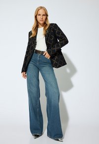 Blazer negro con patrón geométrico dorado, camiseta blanca, jeans de pierna ancha azul de cintura alta, cinturón negro, botas plateadas de punta.