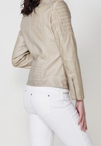Beżowa skórzana kurtka z teksturowanymi akcentami na ramionach i zamkami błyskawicznymi, noszona z białymi dżinsami. Brak widocznych logo lub wzorów.