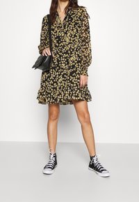 Abito nero floreale con maniche lunghe e orlo a volant, con fiori gialli; abbinato a sneakers alte nere e una borsa a tracolla nera.