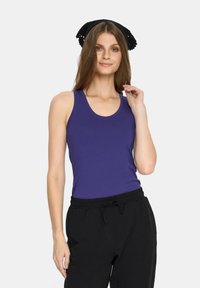 Fioletowy top bez rękawów z gładkiego materiału, w po�łączeniu z czarnymi spodniami na sznurki, modelka delikatnie dotykająca swojej szyi, neutralne tło.