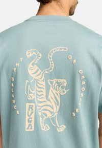 Lichtblauw katoenen T-shirt met een crèmekleurige tijgerafbeelding op de achterkant, vergezeld van de tekst "DE BALANS VAN TEGENSPOREN."