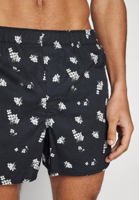 Schwarze Shorts mit weißem Blumendesign, ausgestattet mit einem elastischen Bund. Der Stoff wirkt leicht und hat eine glatte Textur.
