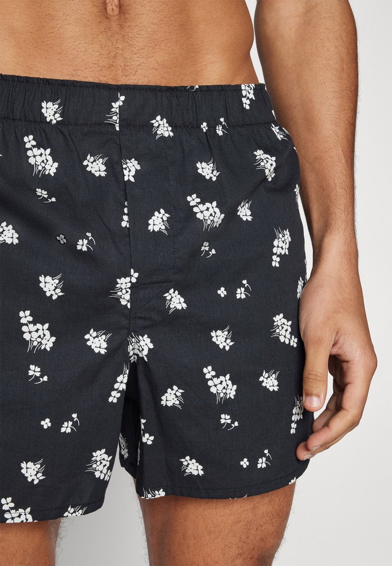 Czarne spodenki z białym wzorem kwiatowym, z elastycznym paskiem w talii. Materiał wydaje się lekki i gładki w dotyku.