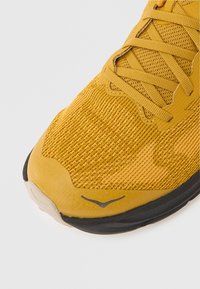 Κίτρινο αθλητικό παπούτσι με πλέγμα και υφή στο επάνω μέρος, επίπεδα κορδόνια και μαύρη λαστιχένια σόλα. Σημαντική λεπτομέρεια branding σε γκρι κοντά στα δάχτυλα.