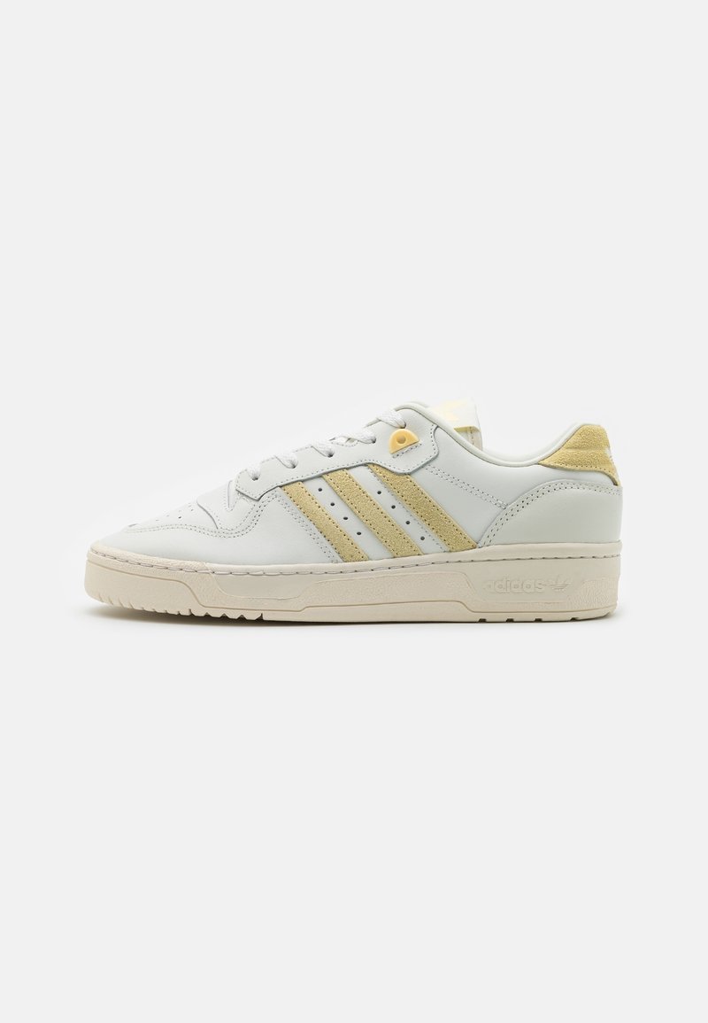 White leather sneakers with light yellow accents and three horizontal stripes. Features a textured sole and rounded toe. Minimalist design.