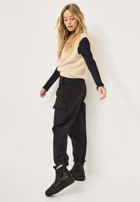 Pull à col roulé beige à côtes sans manches superposé sur une chemise noire à manches longues, associé à un pantalon cargo noir et des bottines noires avec des panneaux élastiques.
